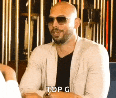 a bald man wearing sunglasses is sitting at a table and says top g .