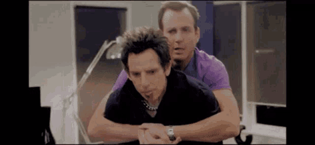 two men are hugging each other in a room . one of the men is wearing a purple shirt .