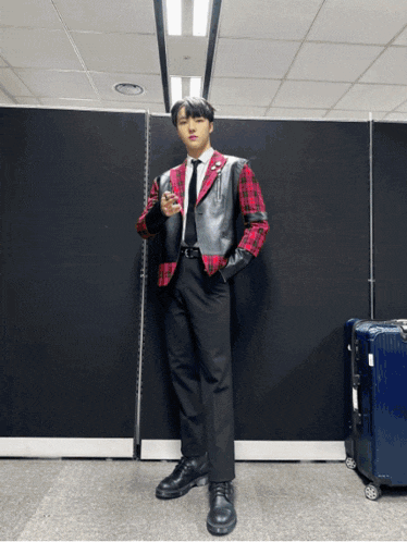 a young man wearing a plaid jacket and black pants stands in front of a blue suitcase