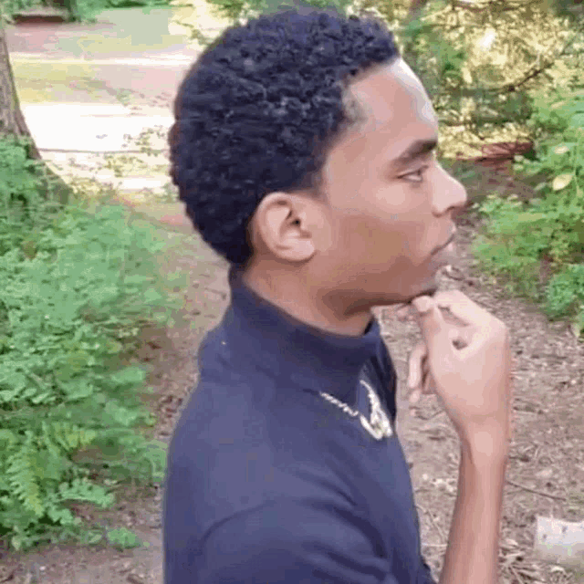 a young man with curly hair is standing in the woods with his hand to his chin .