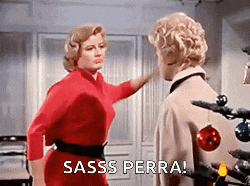a woman in a red dress is pointing at another woman in front of a christmas tree and the woman is saying sass perra