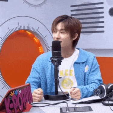 a young man in a blue cardigan is sitting in front of a microphone with a k-pop sign in front of him