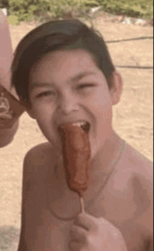 a shirtless young boy is eating a corn dog on a stick