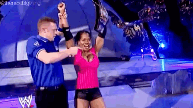 a woman in a pink top is holding a wrestling championship while a man in a blue shirt holds her hands up .