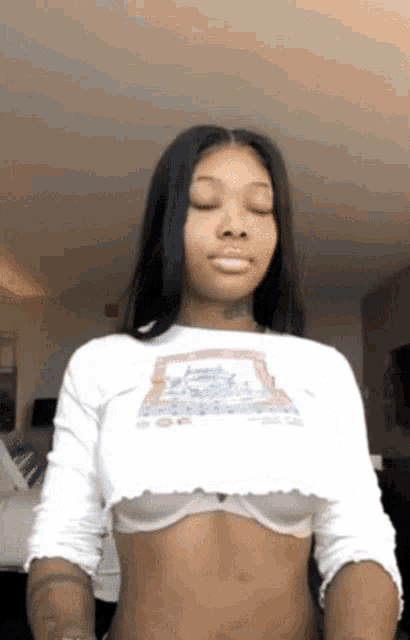 a woman in a white crop top and bra is standing in a room with her eyes closed .