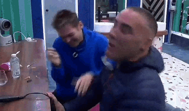 two men are sitting at a table and one of them is wearing a blue sweatshirt