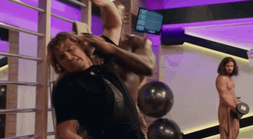 a man is stretching his arms in a gym while another man holds a dumbbell