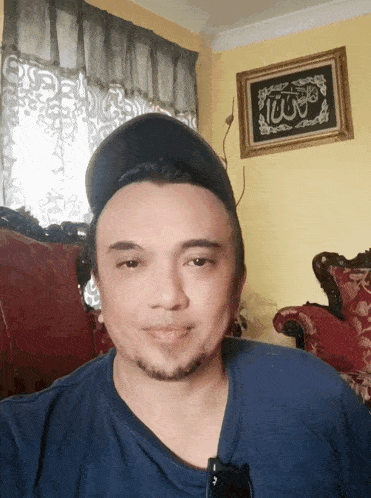 a man wearing a hat and a blue shirt is sitting in front of a framed picture that says ' allah ' on it