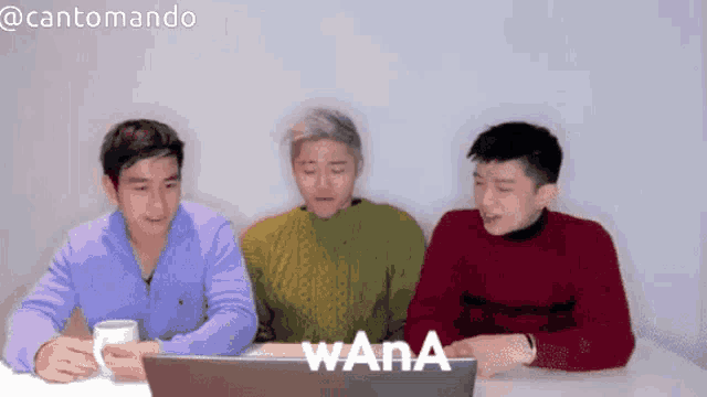 three young men are sitting at a table looking at a laptop screen that says wana on it