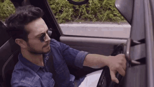a man wearing sunglasses is driving a car with his hand on the steering wheel