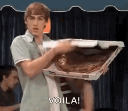 a man is holding a large pizza in a box and says voila .