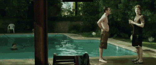 a man standing next to a swimming pool talking to another man with a drink in his hand