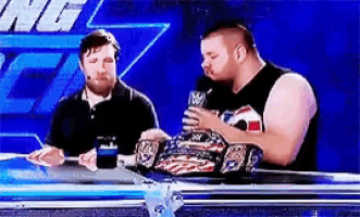 two men are sitting at a table talking to each other while one of them is holding a wrestling championship belt .