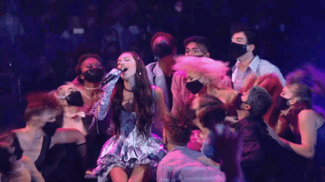 a woman singing into a microphone in front of a crowd wearing face masks