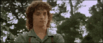 a young man with curly hair is standing in front of trees looking at something .