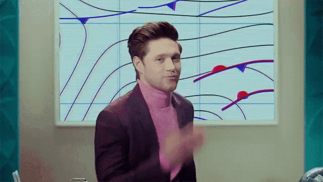 a man in a suit and pink turtleneck is standing in front of a wall with a painting on it .