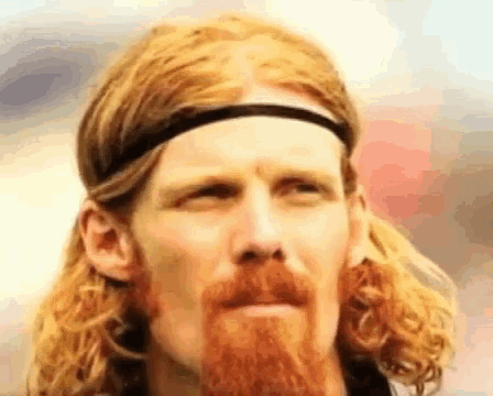 a man with long red hair and a beard wears a headband