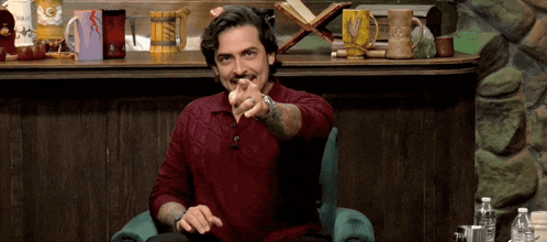 a man in a red sweater is sitting in a chair pointing at the camera .