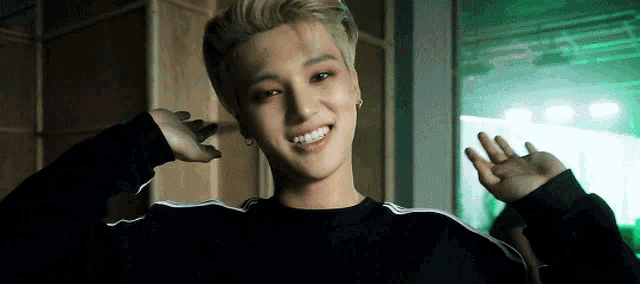 a young man with blonde hair is wearing a black sweater and waving his hand .