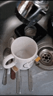 a dirty coffee mug sits in a kitchen sink surrounded by spoons and knives