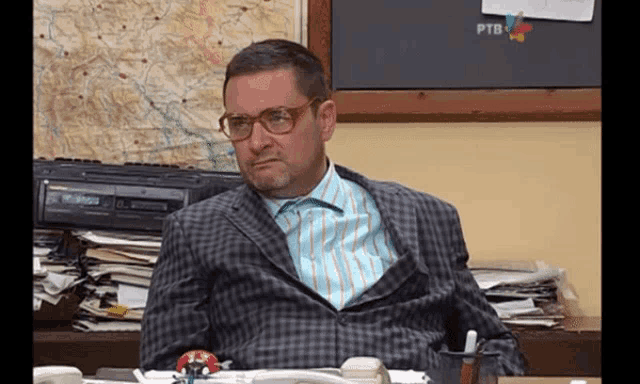 a man wearing glasses and a suit sits at a desk in front of a ptb sign