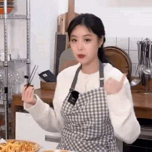 a woman is wearing an apron and holding chopsticks in a kitchen .
