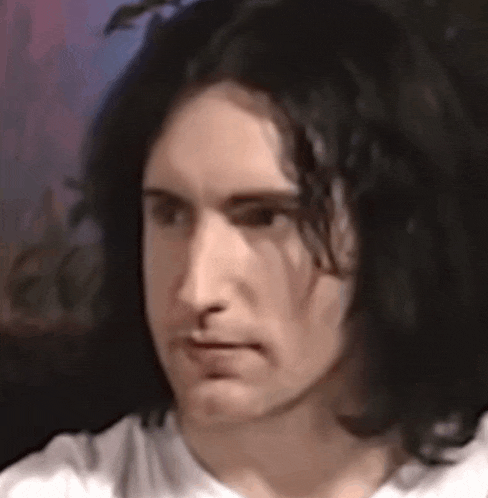 a man with long black hair is wearing a white shirt and making a face .