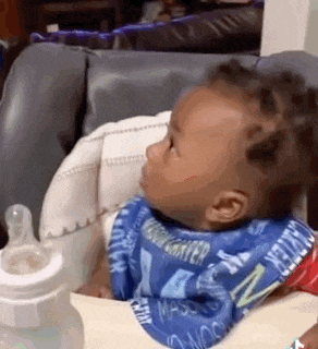 a baby wearing a bib is sitting in a high chair next to a bottle of milk .