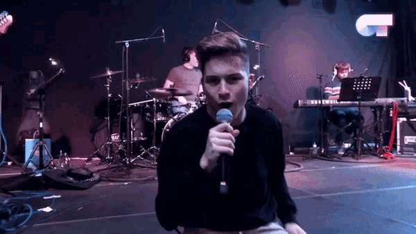 a man singing into a microphone on a stage in front of a drum set