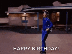 a man in a blue suit is dancing in front of a building and says happy birthday .