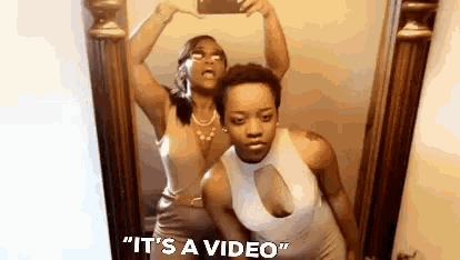 two women are taking a selfie in front of a mirror with the words `` it 's a video '' written below them .