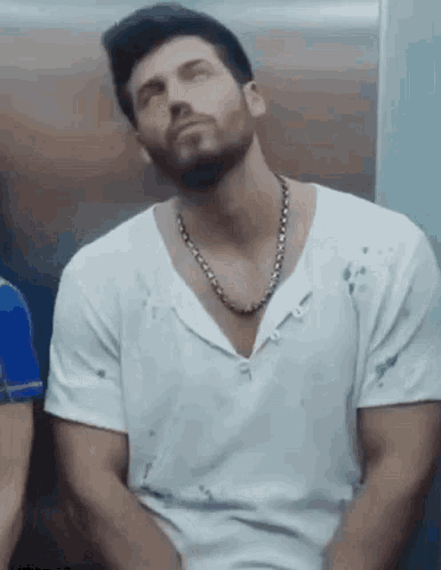 a man with a beard is sitting in an elevator wearing a white shirt and a necklace .