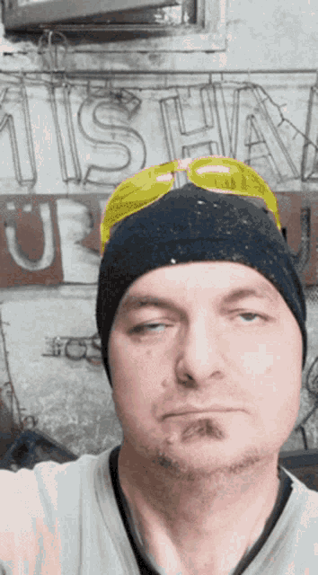 a man wearing a beanie and yellow goggles looks at the camera in front of a sign that says misha