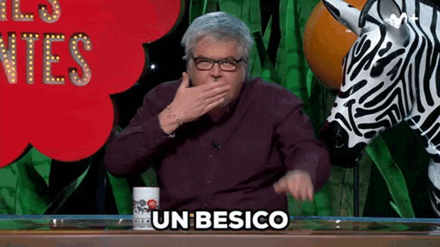 a man with glasses is covering his mouth with his hand and the word un besico is on the table in front of him