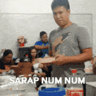 a man in a gray shirt is holding a box in front of a group of people with the caption sarap num num