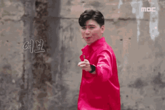 a man in a red shirt is pointing at the camera while standing in front of a wall .