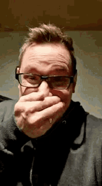 a man wearing glasses is covering his mouth with his hand while making a funny face .
