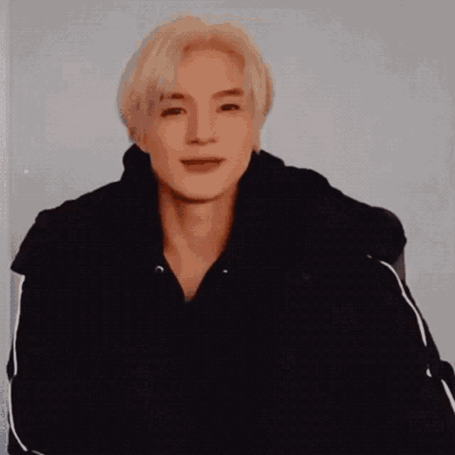 a man with blonde hair is wearing a black jacket and looking at the camera .