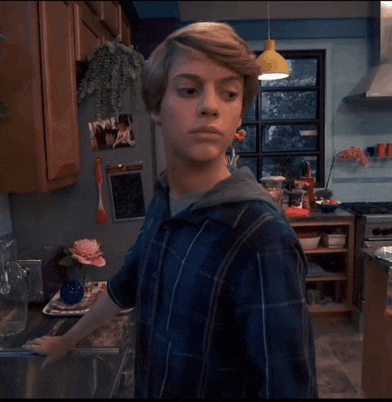 a young man in a blue plaid shirt stands in a kitchen