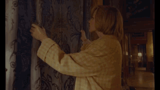 a woman in a yellow coat is holding a curtain in a room