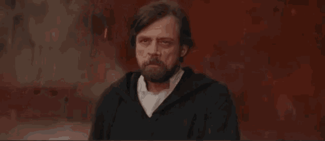 a man with a beard is sitting at a table with his hands folded in front of him .