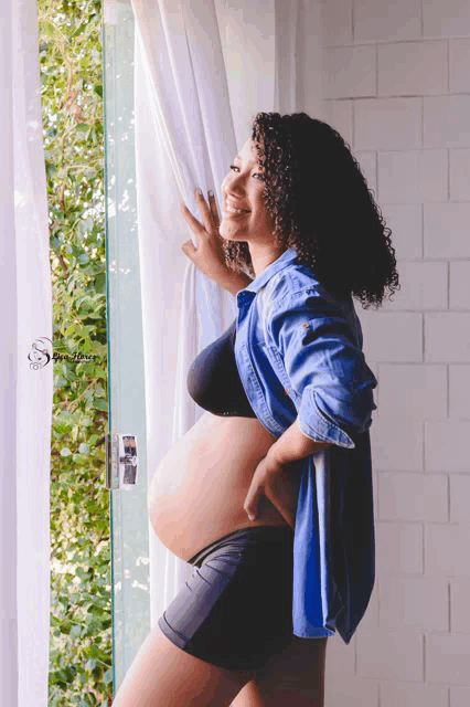 a pregnant woman in a blue shirt and black shorts looks out a window and smiles