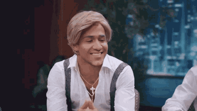 a man wearing a wig and a necklace is smiling while sitting on a chair .