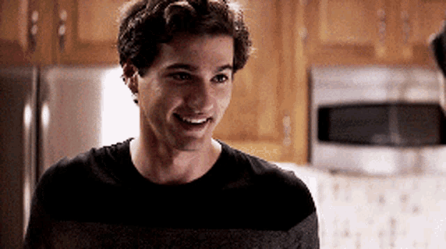 a young man is smiling in a kitchen with a refrigerator and microwave in the background .