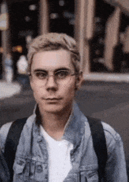 a young man wearing glasses and a denim jacket is standing in front of a building .