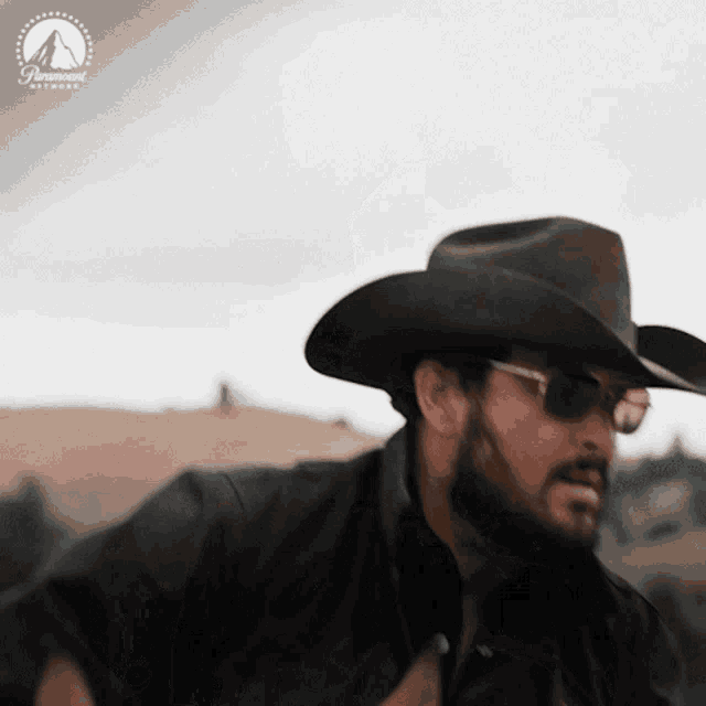 a man wearing a cowboy hat and sunglasses is standing in front of a mountain .