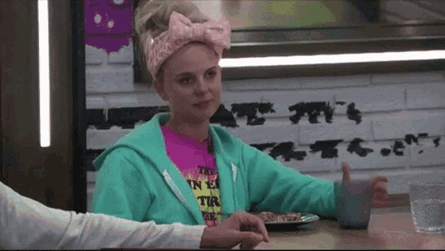 a woman wearing a pink headband and a green hoodie sits at a table with a plate of food ..