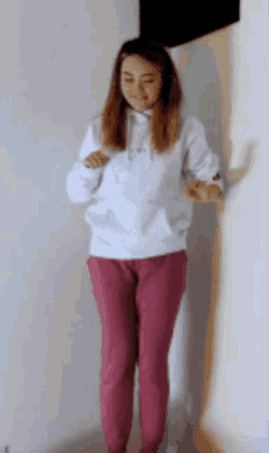 a woman in a white hoodie and pink sweatpants is standing in front of a white wall .