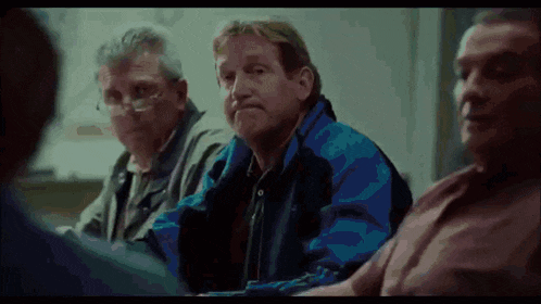 a group of men are sitting around a table and one of them is wearing a blue jacket .