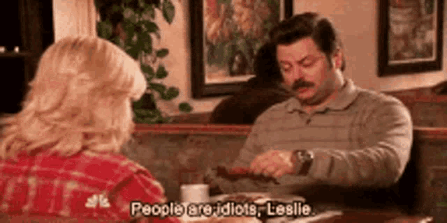 a man and a woman are sitting at a table in a restaurant and the man is saying people are idiots leslie .